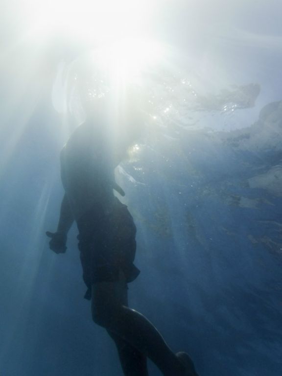 An underwater photograph looking up towards a young swimmer who is surfacing at the point where the sun hits the water.