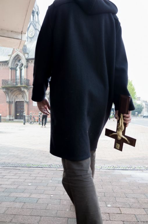 The portrait format image shows the back of a man, cropped at the neck and ankle and dressed in a knee length black, monk-like coat. In his right hand he is holding a brown crucifix with a detailed, white effigy of Christ.