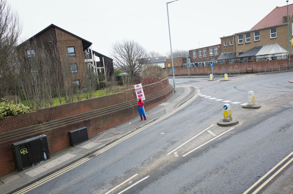 A person dressed in a spiderman outfit stands on a pavement holding a pizza restaurant sign. They take up the middle tenth of the frame and there is no-one driving on the roads or walking on the pavement.