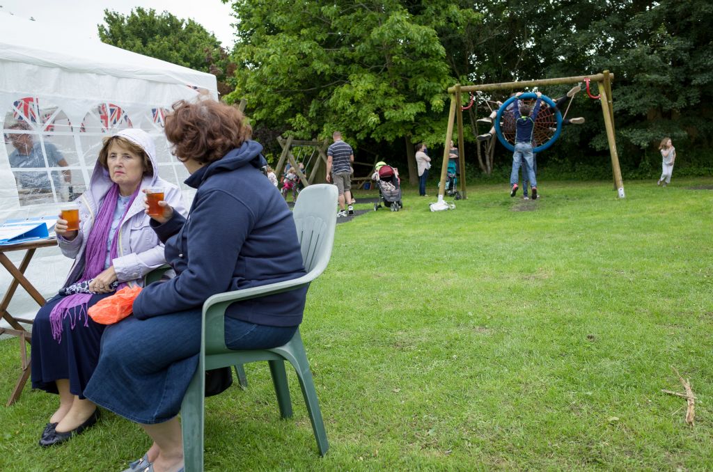 Two women sit on plastic, green seats in left foreground drinking pimms and looking miserable at a traditional English Fete. A group of kids swing at a park in the right background.