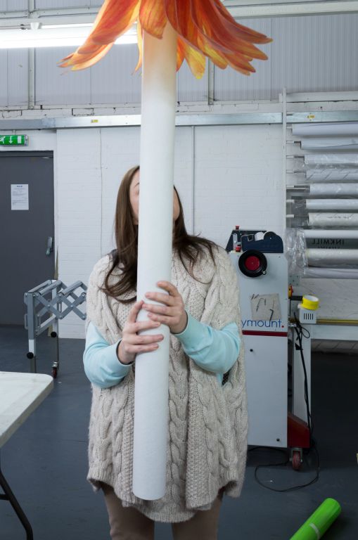 The portrait photo depicts a women holding a white tube wich obscures her face. On top of the tube is a 50 centimetre wide orange, fake flower head.