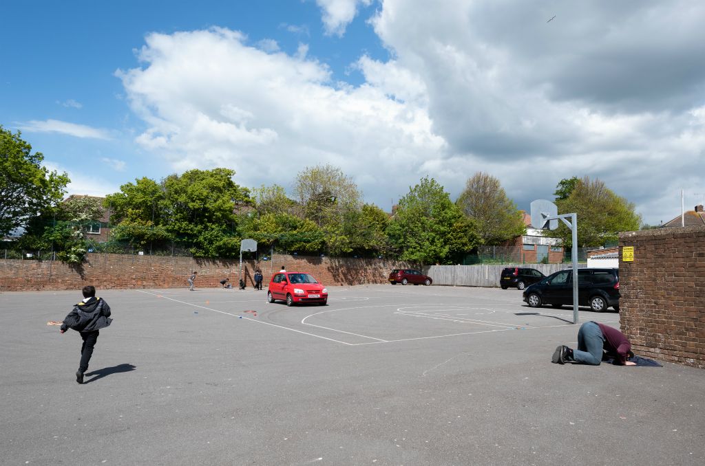 The scene is a playground that is partially being used as a car park with one bright red car in the middle ground. In the Left foreground a boy runs away from the camera towards a game of basketball in the background beyond the red car. In the right foreground a man is kneeling in a praying position with his head on the ground on front of a red brick wall.