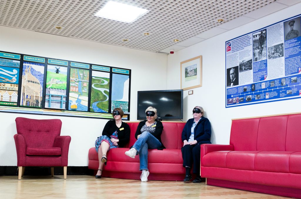 Three ladies with name tags sit on a red sofa with black blindfolds on their eyes.