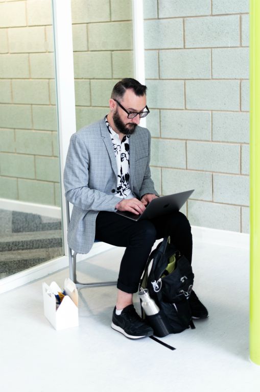 A man sits on a lone chair operating a laptop with a box, presumably lunch, sitting next to him.