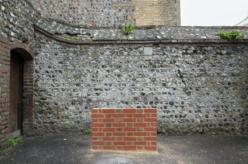 The landscape photograph is filled by an old looking flint wall. About one metre front of the flint wall at filling about one third of the bottom half of the frame is a brand new