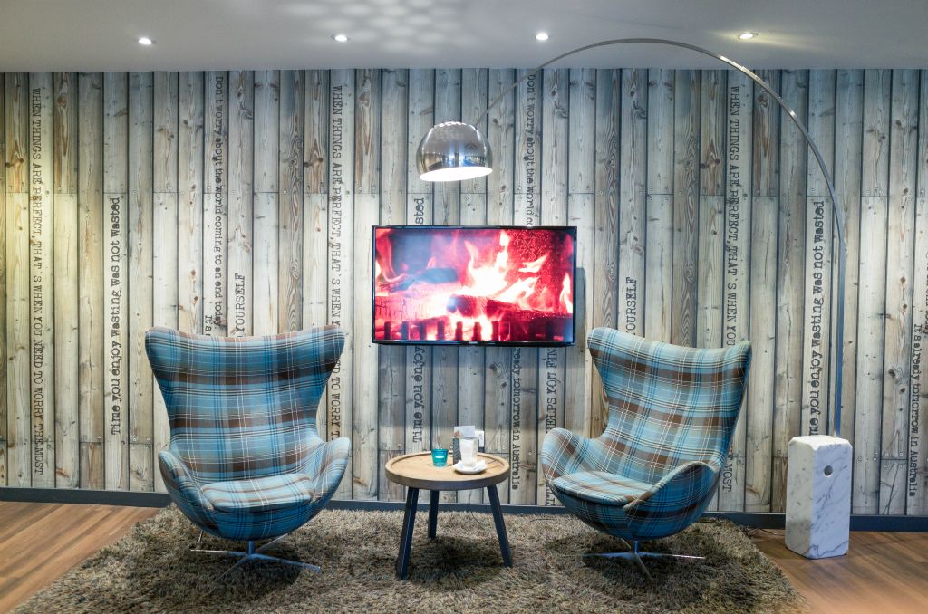 A landscape photo depicts an interir scene with two tartan, comfy, swivel chairs with a small, round table inbetween sitting on a fawn brown, shagpile rug. The centre of the image has a flat screen tv behind the chairs on a walldepicting a burining fire.