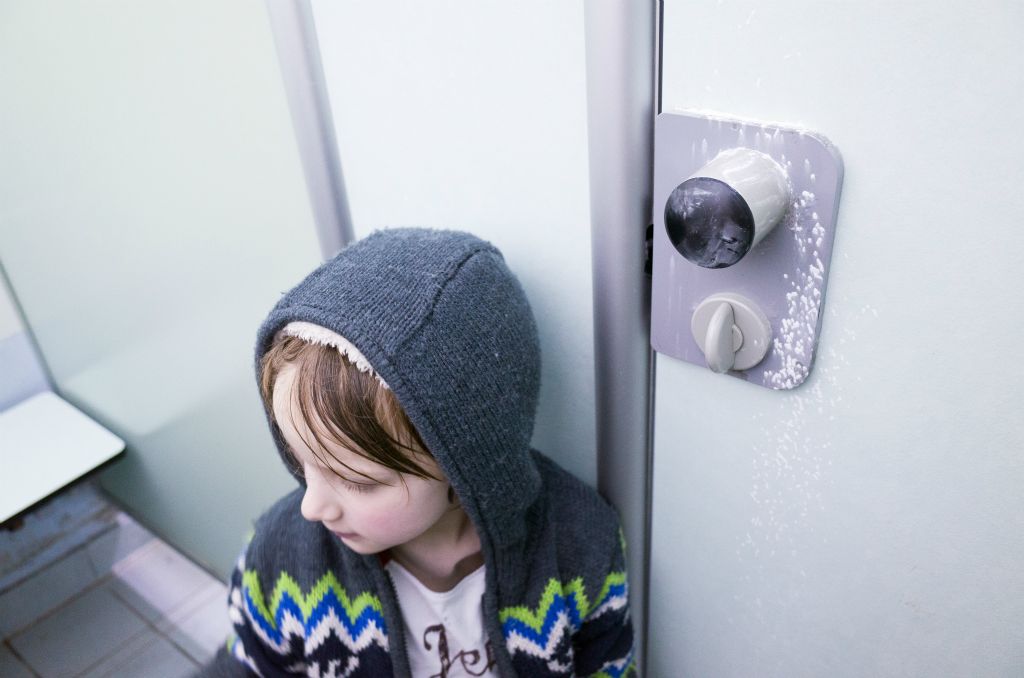 A young boy in hooded, woollen top leans against the wall of a swimming pool cubicle. A badly aimed pepper of talcum powder has hit the door lock and surrounding wall.