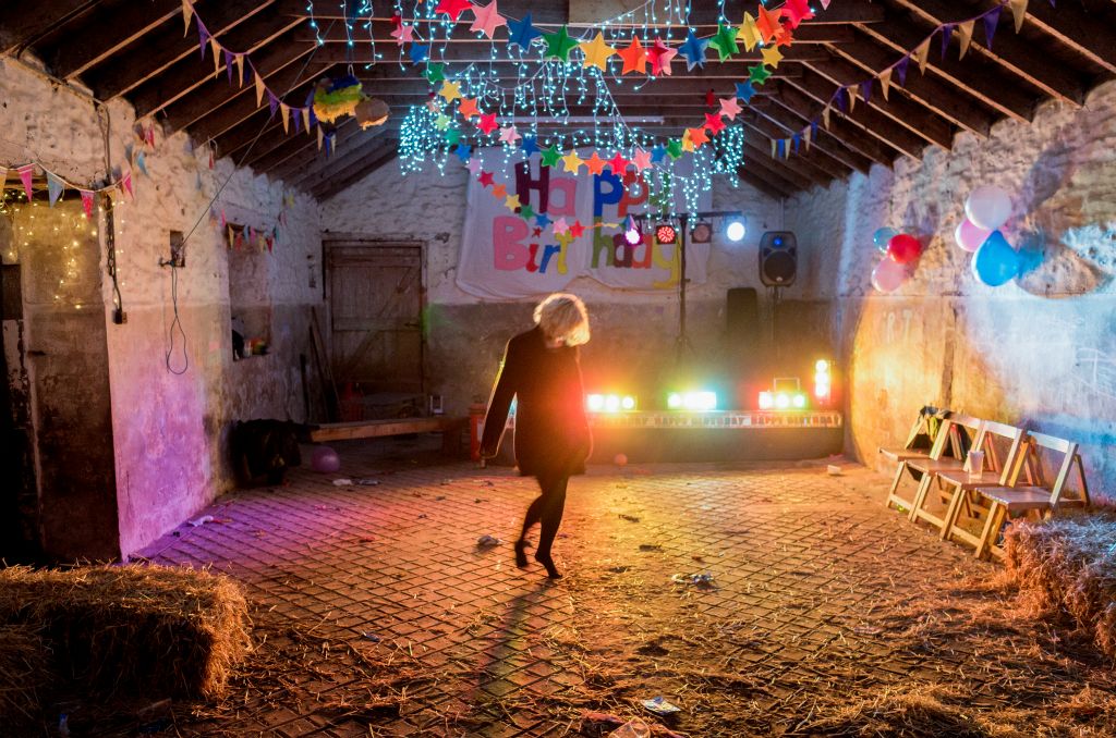 A single female who's face is obscured by her blond hair dances alone in a agricultural shed that's been decorated to celebrate a birthday.