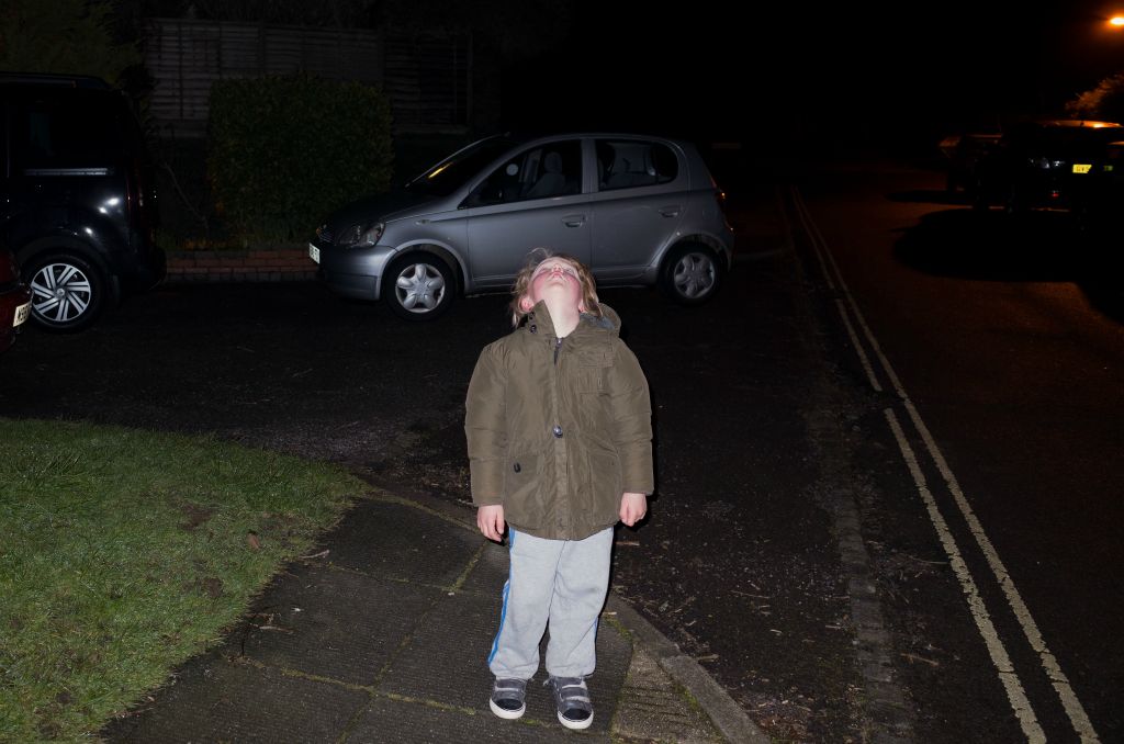 A young boy lit by camera flash at night looking up to the sky.