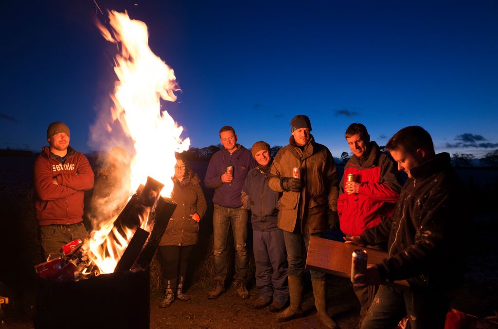 The landscape photograph is dominated by by a tall fire on the left side of the frame. Sevenm indeviduals are lined up curving round the fire against a twilight sky. The indevidual closest to the camera on the right side appears to be reading from a small sheet of wood.